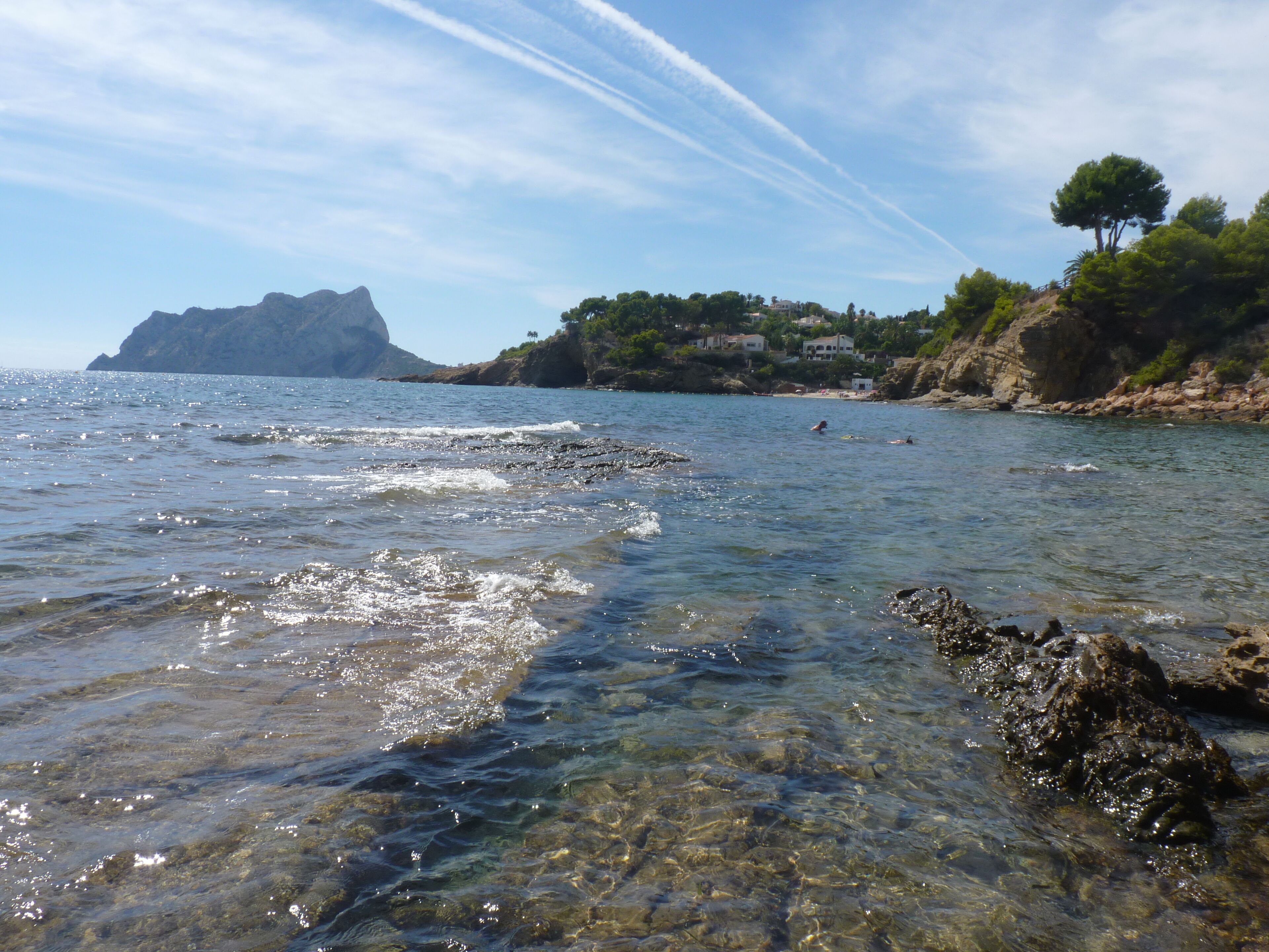 A coastal scene with clear water, rocky shore, and a distant mountain.