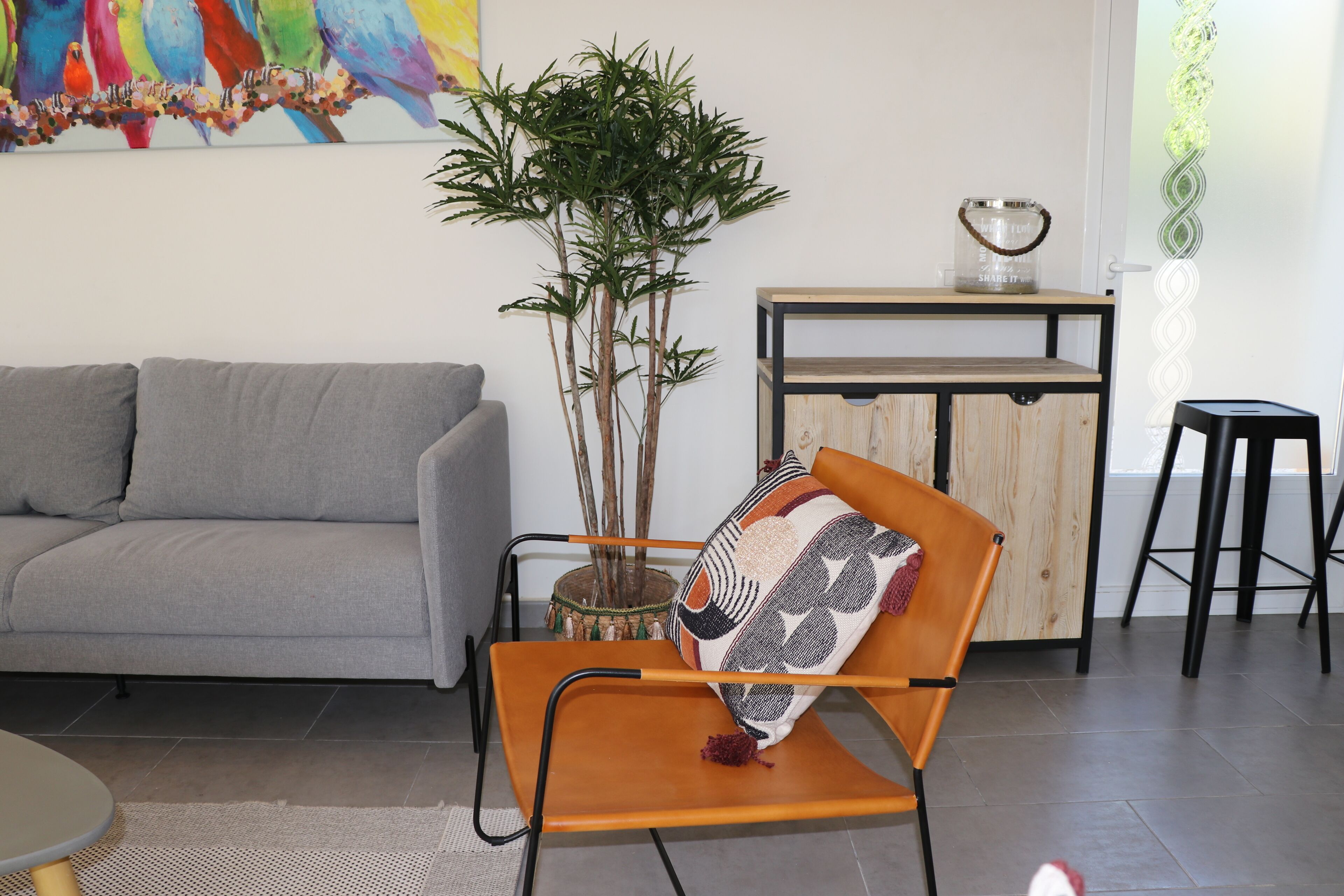 A modern living room with a grey sofa, an orange chair with a patterned cushion, a tall green plant, and a wooden side table with a decorative item.