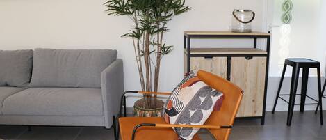 A modern living room with a grey sofa, an orange chair with a patterned cushion, a tall green plant, and a wooden side table with a decorative item.