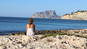 En kvinde sidder på en stenet strand og ser ud mod en klippe med et vandområde i baggrunden.