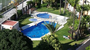 Zona de piscina en la azotea con dos piscinas, rodeada de palmeras y un edificio blanco.