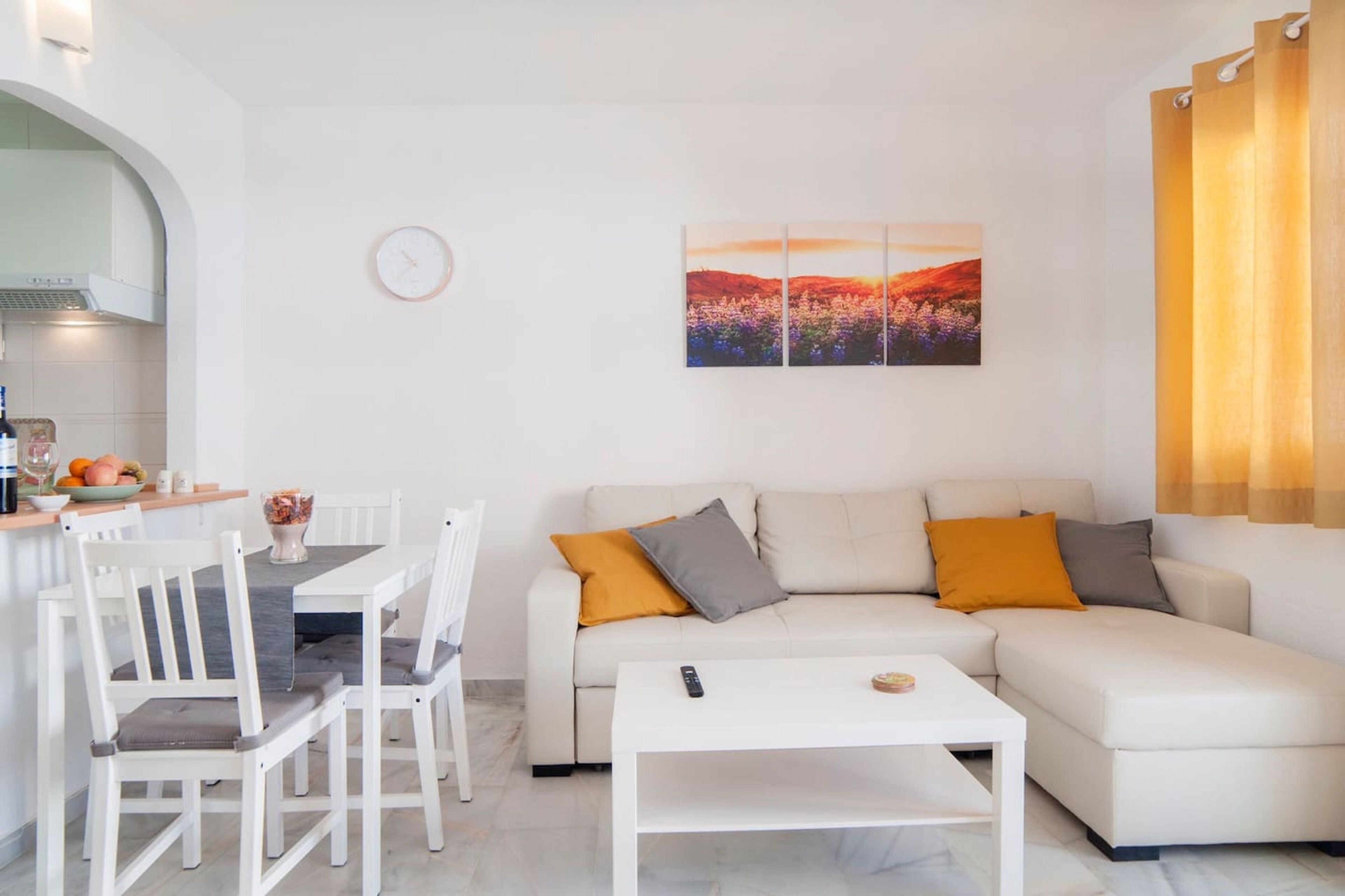 A modern living room with a white sofa, a dining table with chairs, a small white coffee table, and a painting on the wall.