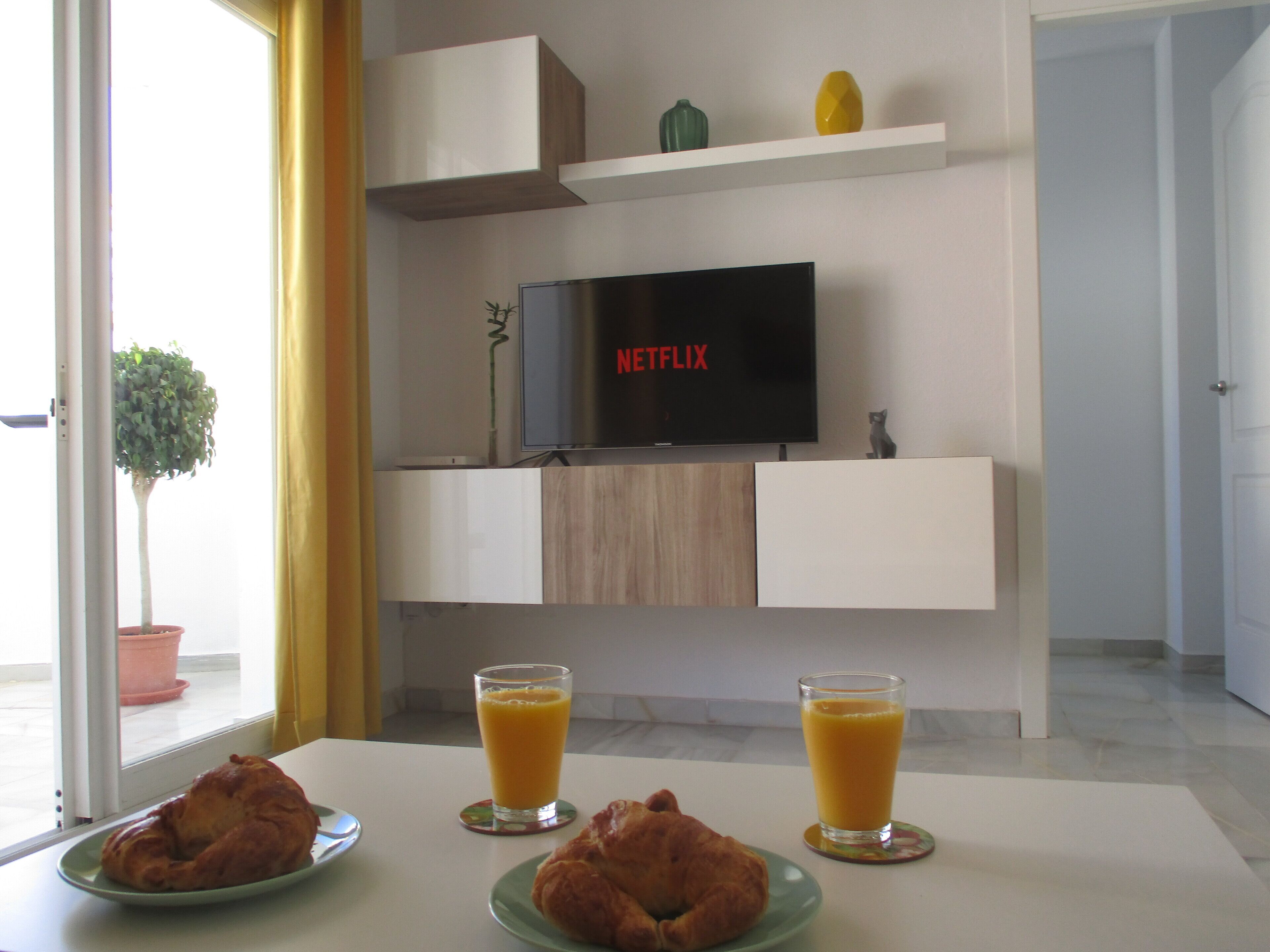 A modern living room with a flat-screen TV displaying 'NETFLIX', a potted plant, and two glasses of orange juice on a table.