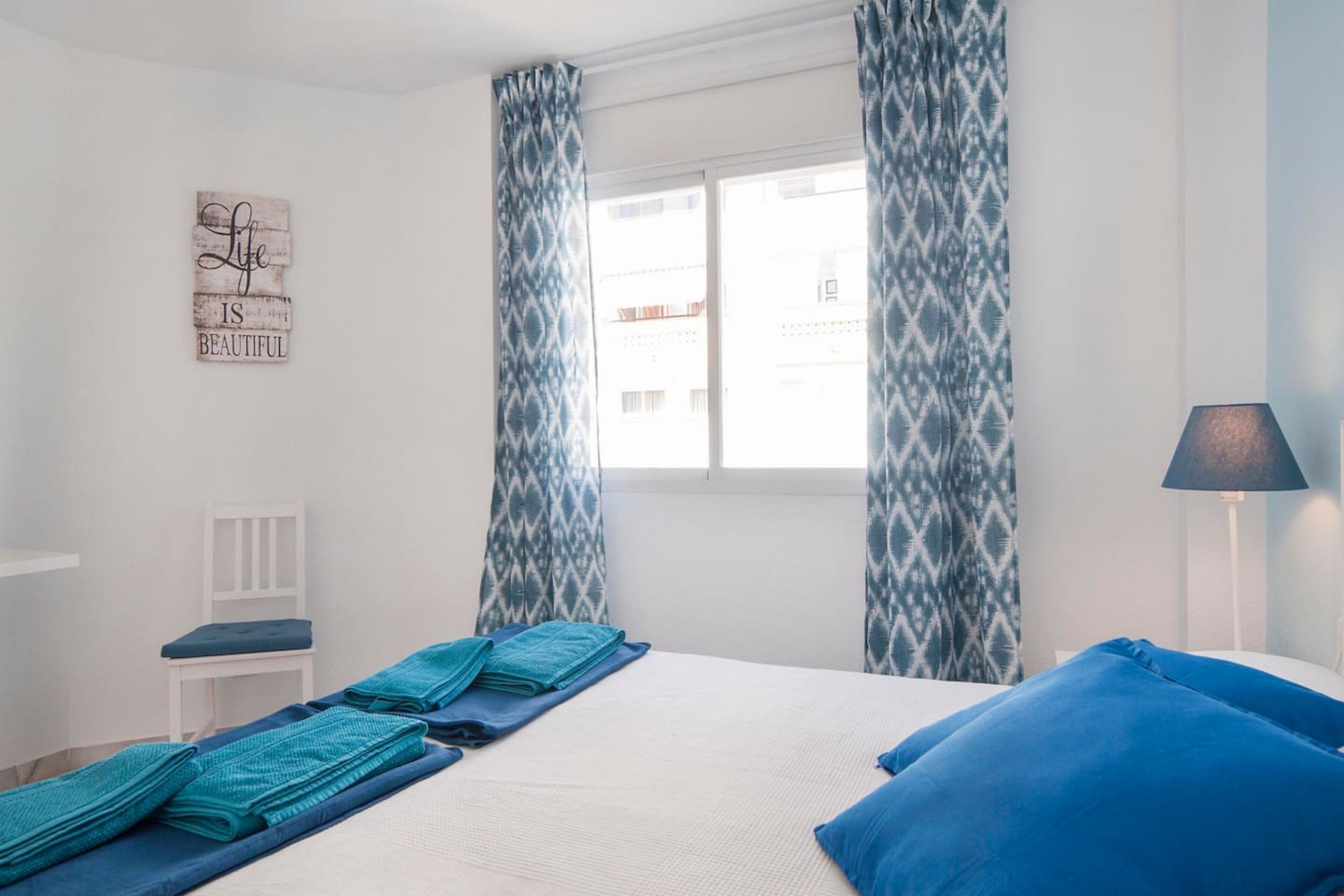 A bedroom with a bed, blue and white patterned curtains, a small white chair, and a wall-mounted sign.