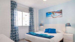 Habitación de hotel con cama, dos mesitas de noche con lámparas, una ventana con cortinas y un cuadro con motivos playeros en la pared.