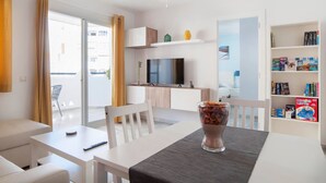 Un salón moderno con televisor de pantalla plana, una mesa de comedor con sillas, una estantería y un balcón con cortinas.