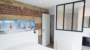 A bedroom with a brick wall, a large painting, a white bed, and a bathroom with a glass shower. - Charming Villa with Garden near the beach (Tourgéville)