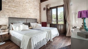 Ein Schlafzimmer mit zwei Betten, einer Steinwand, Holzbalken, einem Fenster mit Vorhängen und einem Nachttisch mit Lampe.