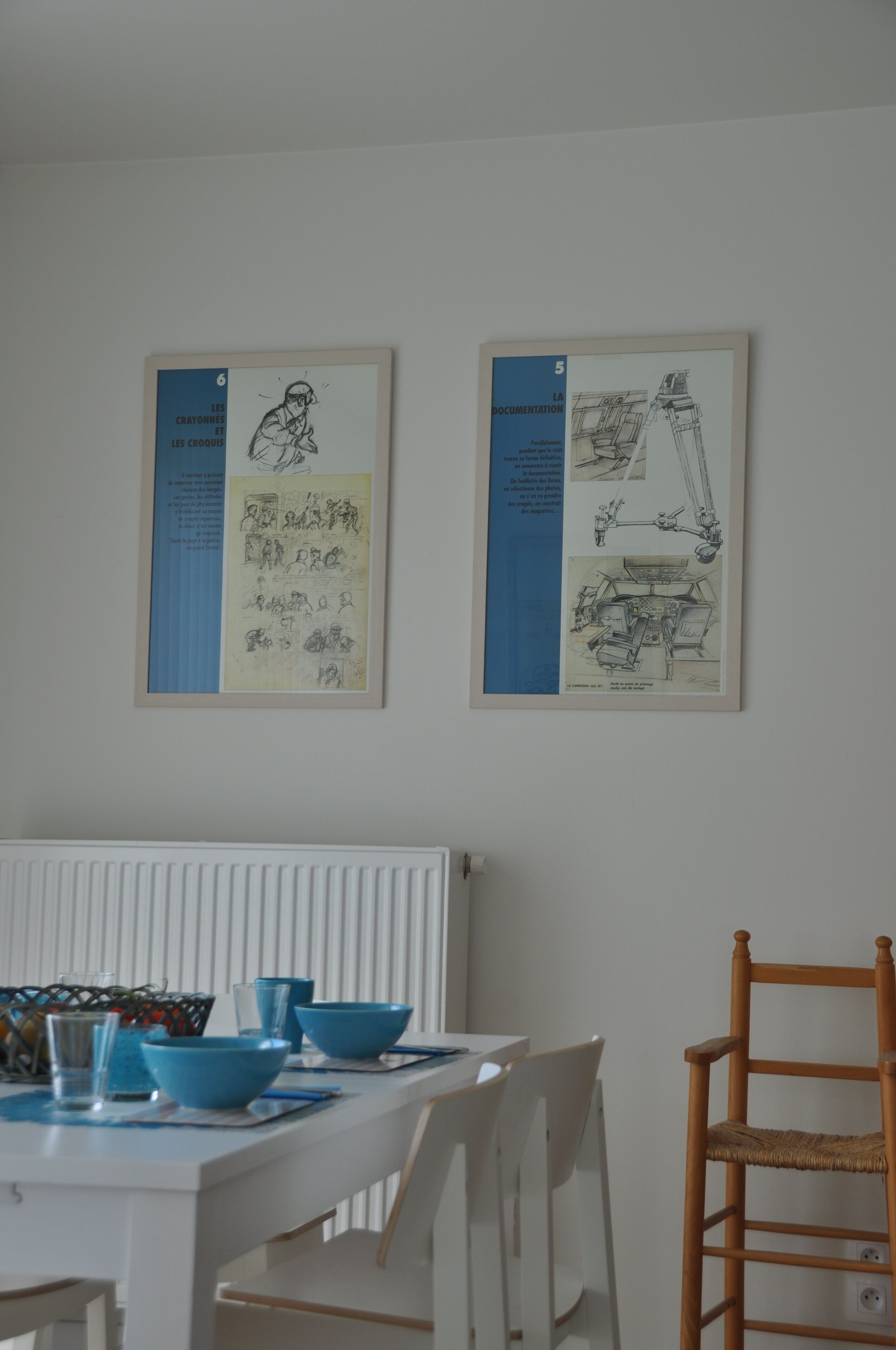 Un comedor con una mesa blanca, cuencos azules y sillas. Hay dos documentos enmarcados en la pared.