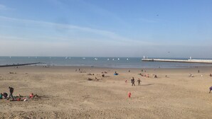 Una extensa playa arenosa con un muelle que se adentra en el agua, varios veleros y gente realizando diversas actividades.