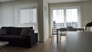 Una sala de estar con un sofá de color oscuro, una mesa de comedor con sillas y ventanas grandes con persianas.