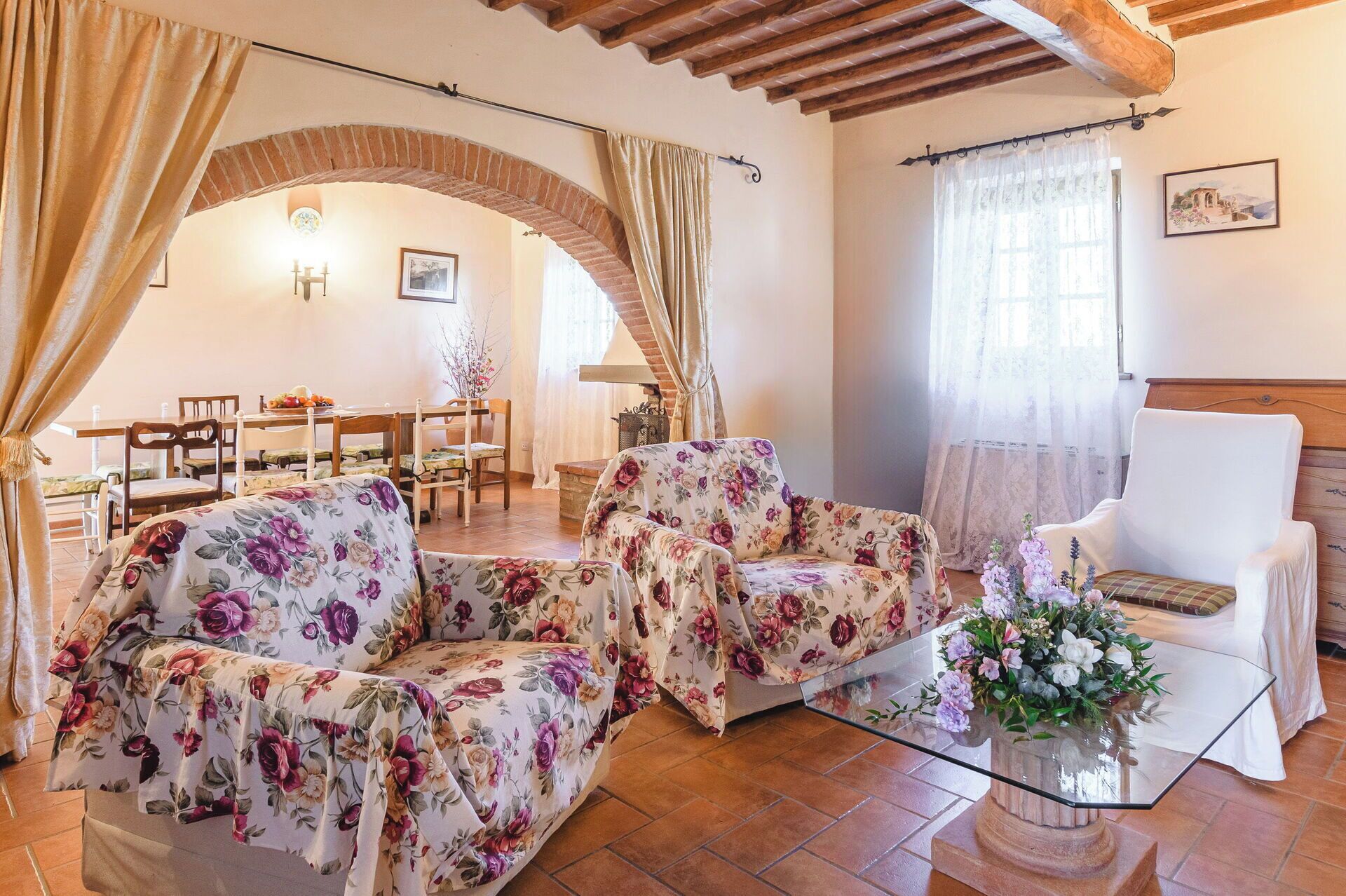 Un salon chaleureux avec des canapés à motifs floraux, une table basse en verre et un fauteuil blanc. Un arc donne accès à un coin repas avec une table et des chaises.