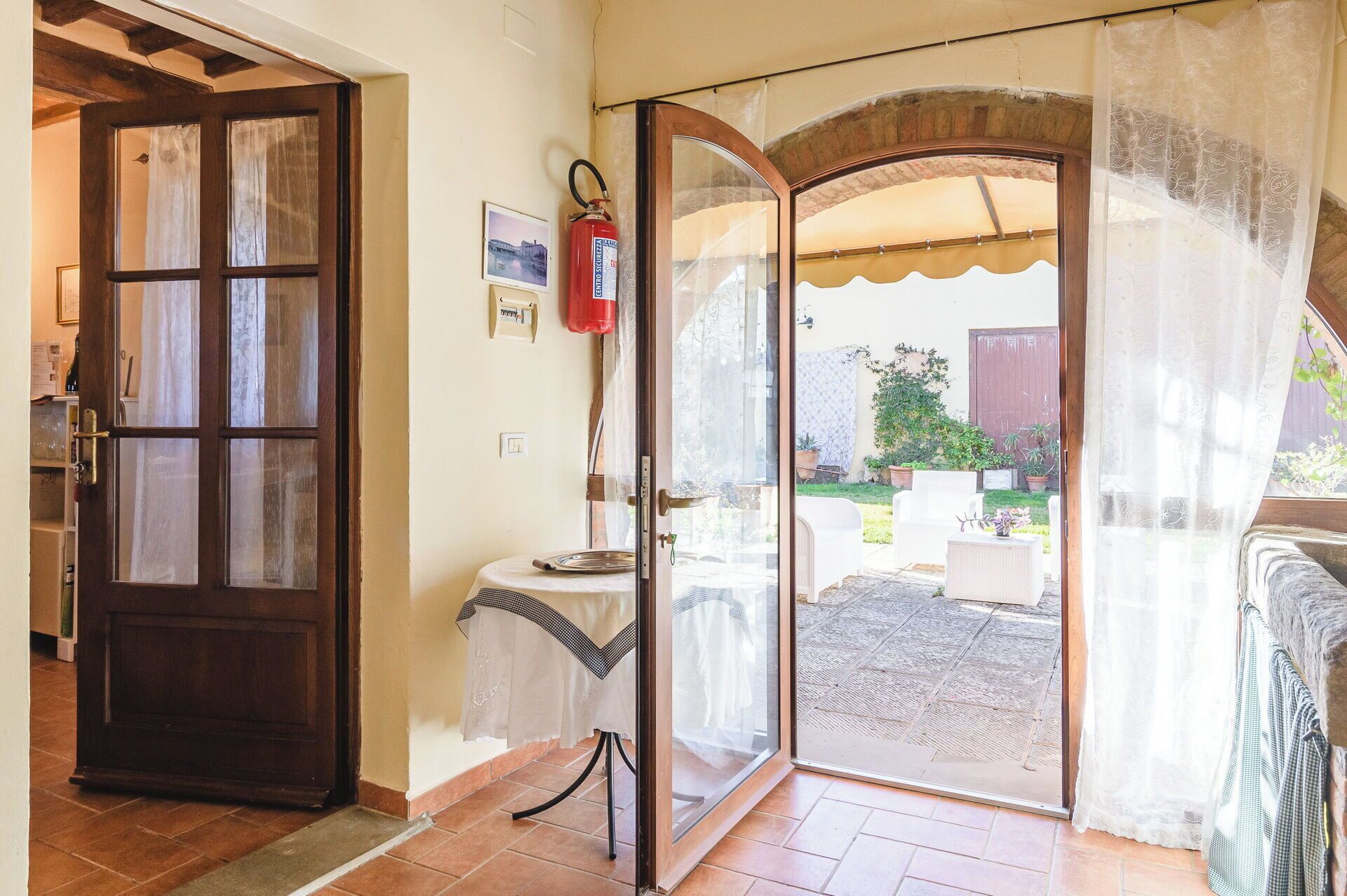 Une pièce avec une porte en bois, une petite table recouverte d’une nappe à carreaux et une vue sur un patio extérieur aménagé avec une table et des chaises.
