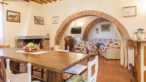 A dining area with a wooden table and chairs, a fireplace, and a bed with floral bedding under a brick archway.