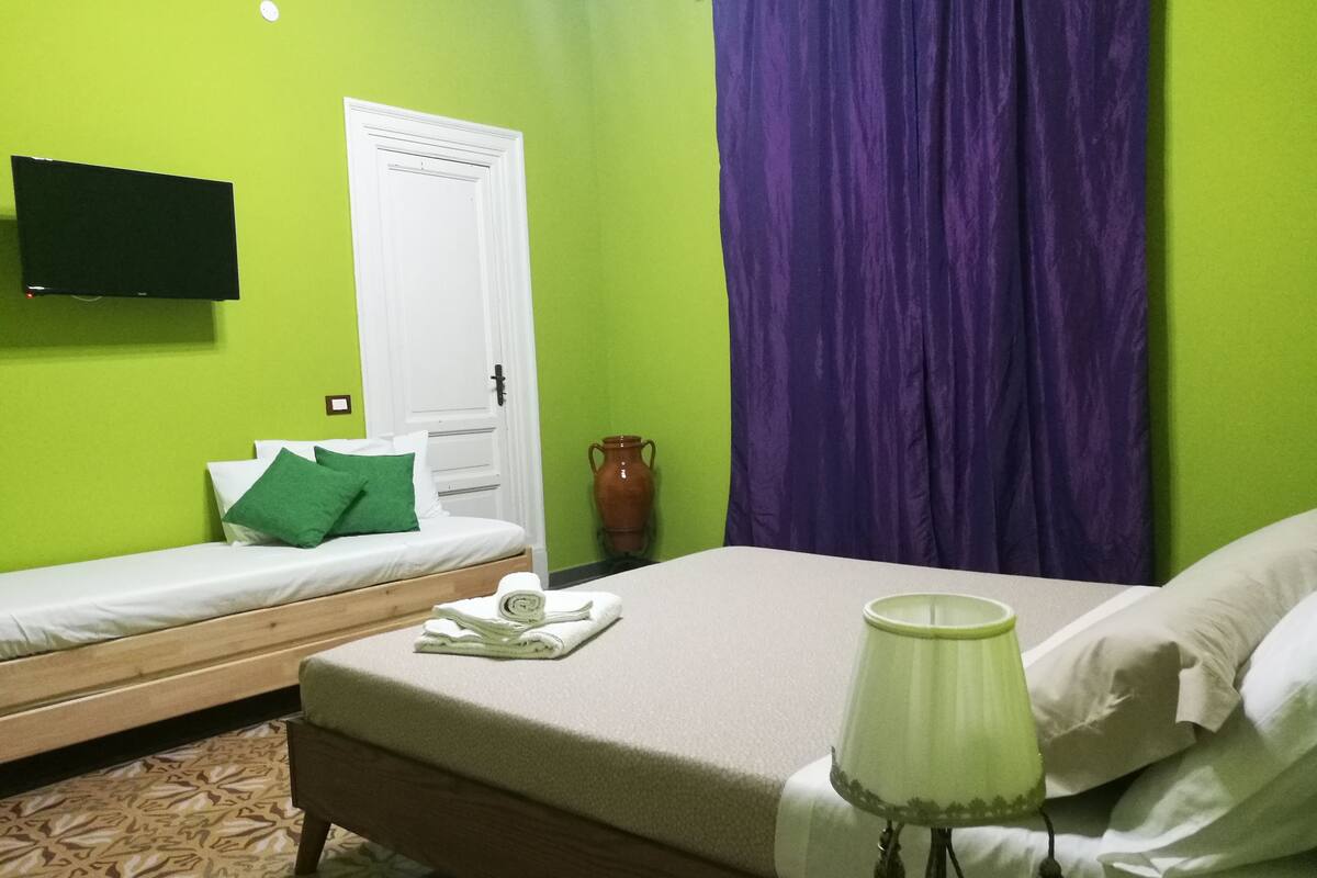 Ein Zimmer mit zwei Betten, einem Fernseher an der Wand und einer Lampe auf dem größeren Bett.