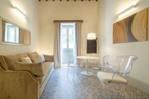 A room with a beige sofa, clear chairs, a round table, a lamp, and a patterned floor.