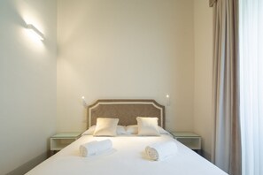 A neatly made bed with pillows and rolled towels, a bedside table, and a wall-mounted light fixture.