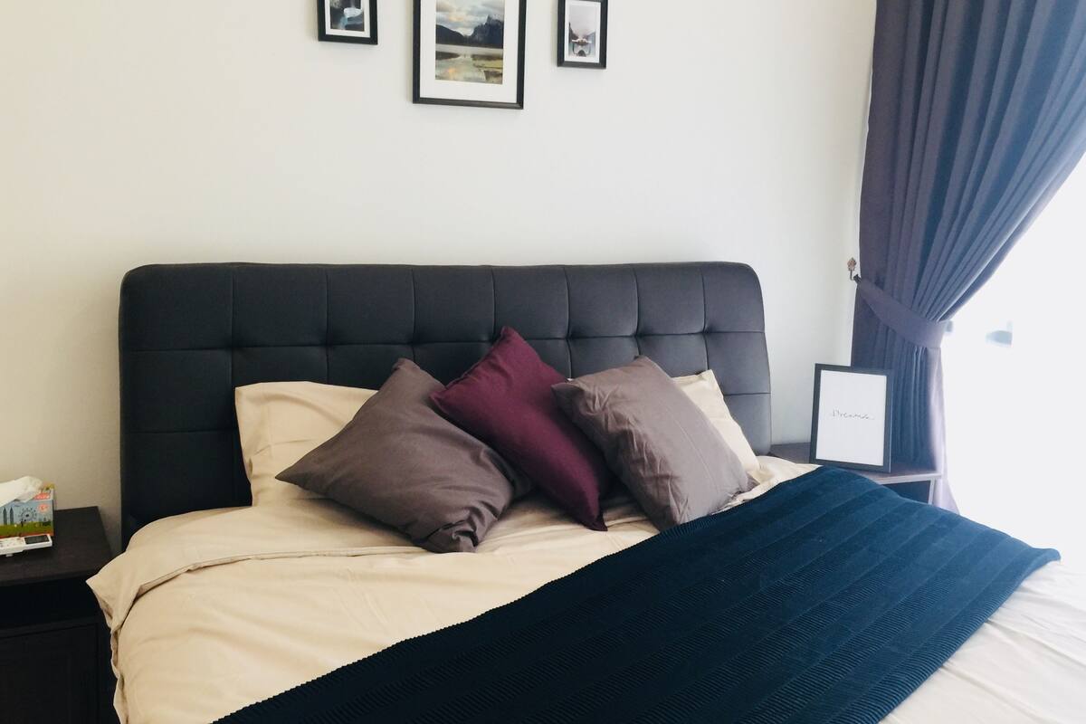En snyggt bäddad säng med en mörk sänggavel, flera kuddar och ett mörkt täcke. På väggen ovanför sängen hänger inramade tavlor.