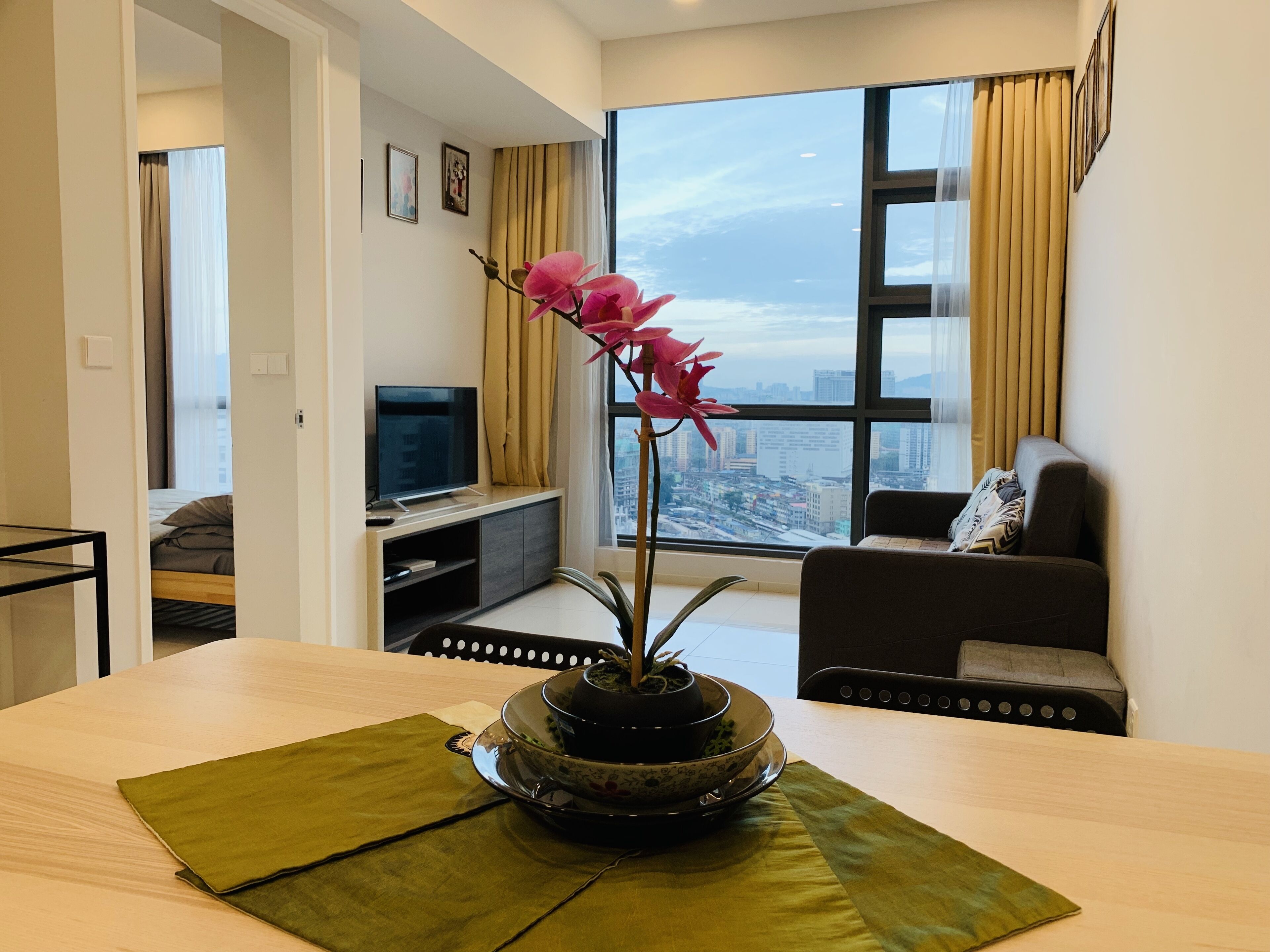 Una sala de estar moderna con un ventanal grande, un televisor de pantalla plana, un sofá y una mesa de comedor con un centro de mesa decorativo con una orquídea.