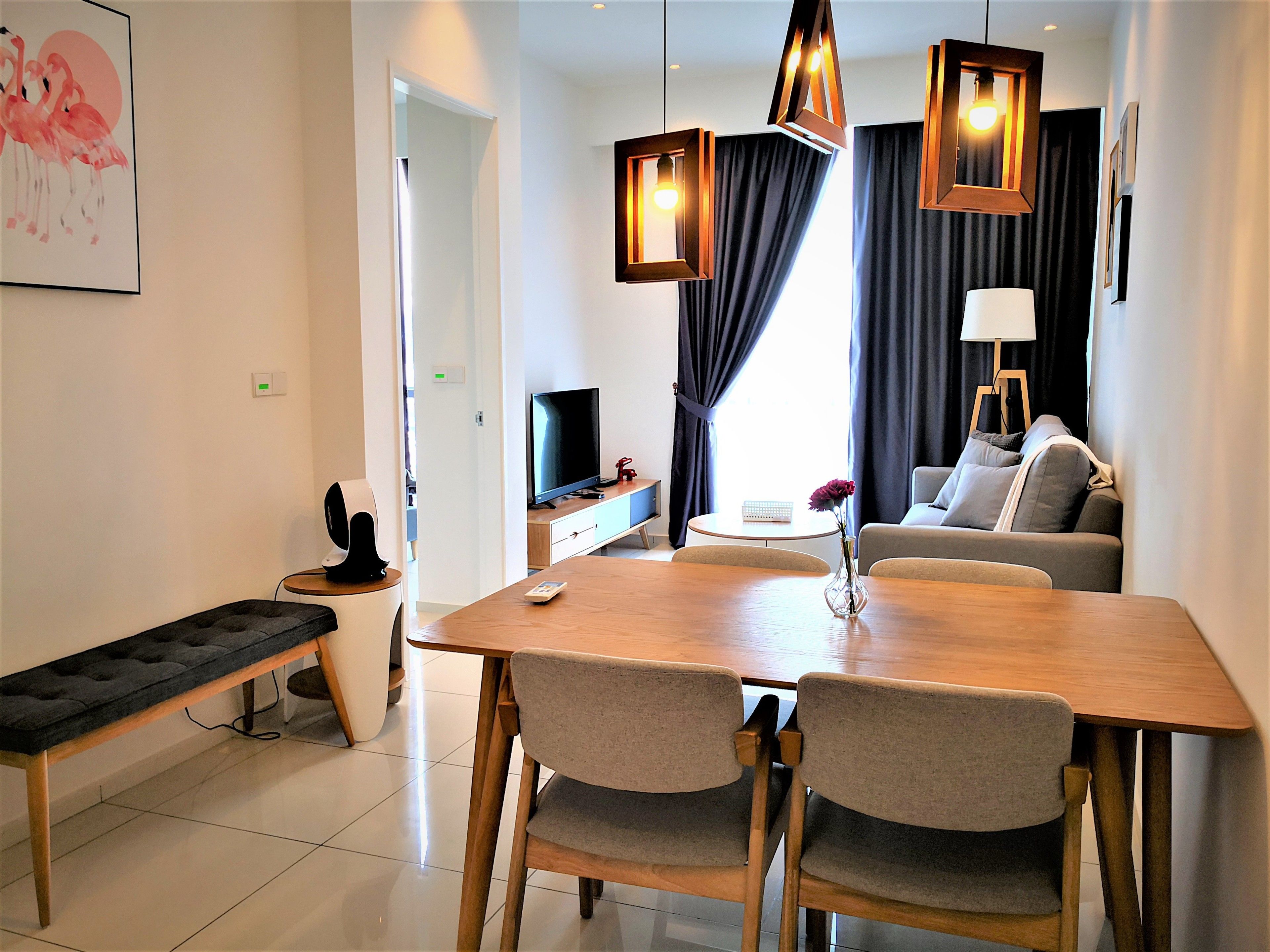Uma sala de estar moderna com uma mesa de jantar de madeira, duas cadeiras estofadas cinzentas, um banco, uma TV de tela plana, uma mesa pequena branca e um sofá com almofadas cinzentas.