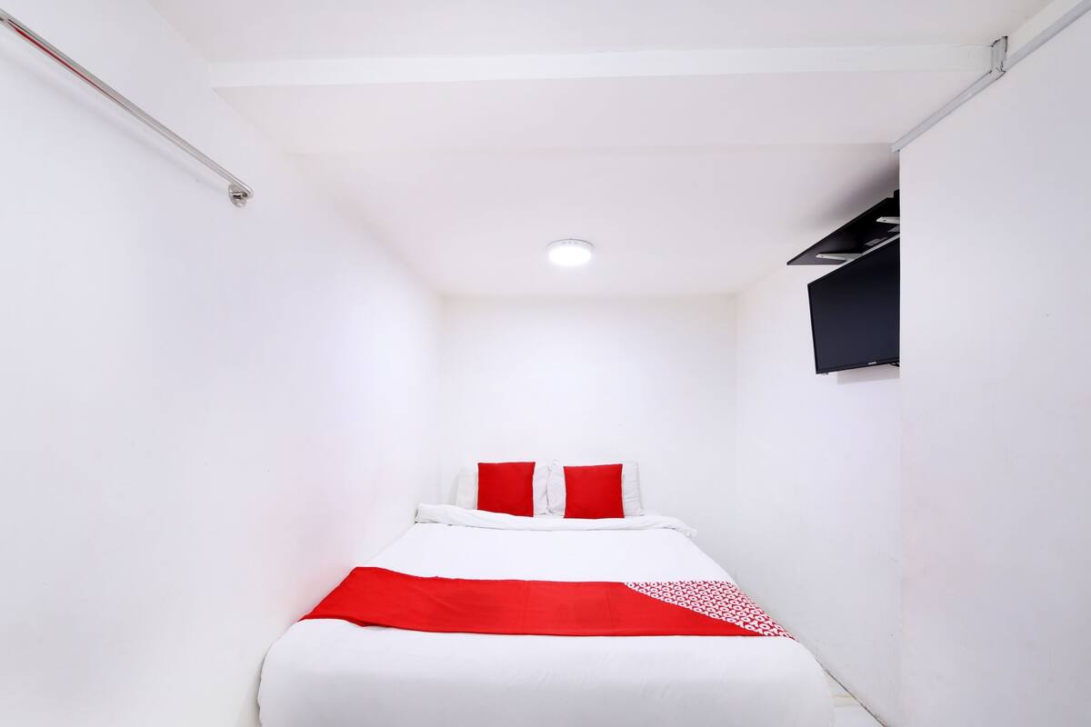Een smalle hotelkamer met een bed, witte muren, een rode en witte gestreepte plaid en een zwart hoofdeinde.