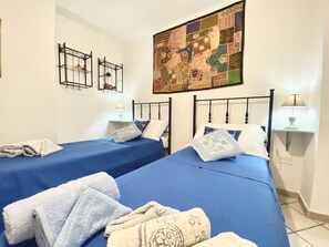 Ein Hotelzimmer mit zwei Betten, jeweils mit blauer Bettdecke und weißen Kissen. An beiden Seiten der Betten befinden sich Wandlampen. Darüber an der Wand hängt eine große, farbenfrohe Stegdecke.