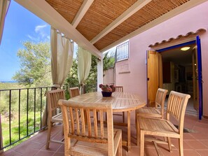 Eine überdachte Terrasse mit einem runden Holztisch und Stühlen, einem Blumentopf und Blick aufs Meer.