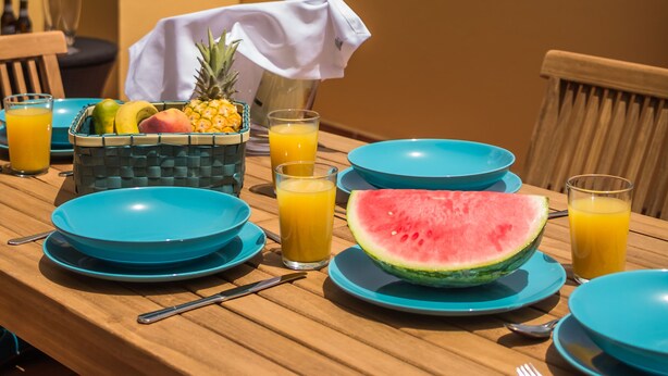 Une table dressée avec des assiettes bleues, une tranche de pastèque et des verres de jus d’orange, donnant sur l’océan.