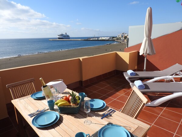 A dining table set with blue plates, cutlery, and a fruit basket, overlooking a beach with a cruise ship in the distance. - Ocean View Penthouse II. Beachfront Wifi & A / C. 23m2 private terrace. (Gran Tarajal)