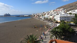 Une plage avec un navire de croisière en arrière-plan, une promenade bordée de boutiques et de palmiers, et une colline avec des bâtiments.