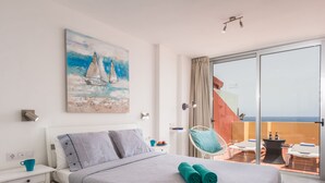 Une chambre avec un lit, un tableau représentant un voilier, un balcon avec une chaise et une table, et une vue sur la mer.