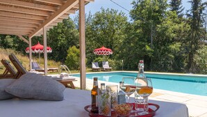 Een zwembadterras met ligbedden, een tafel met drankjes en een pergola.