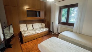 A room with a wooden bed, a sofa bed with patterned cushions, a small table, and a window with curtains. - Apartment in villa with garden CIN IT032003C2BP5JM2B2 (Muggia (TS))
