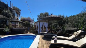 A wooden deck with a swimming pool, lounge chairs, and a thatched-roof structure. - Luxury, self contained, airconditioned studio with private terrace & use of pool (Begur)