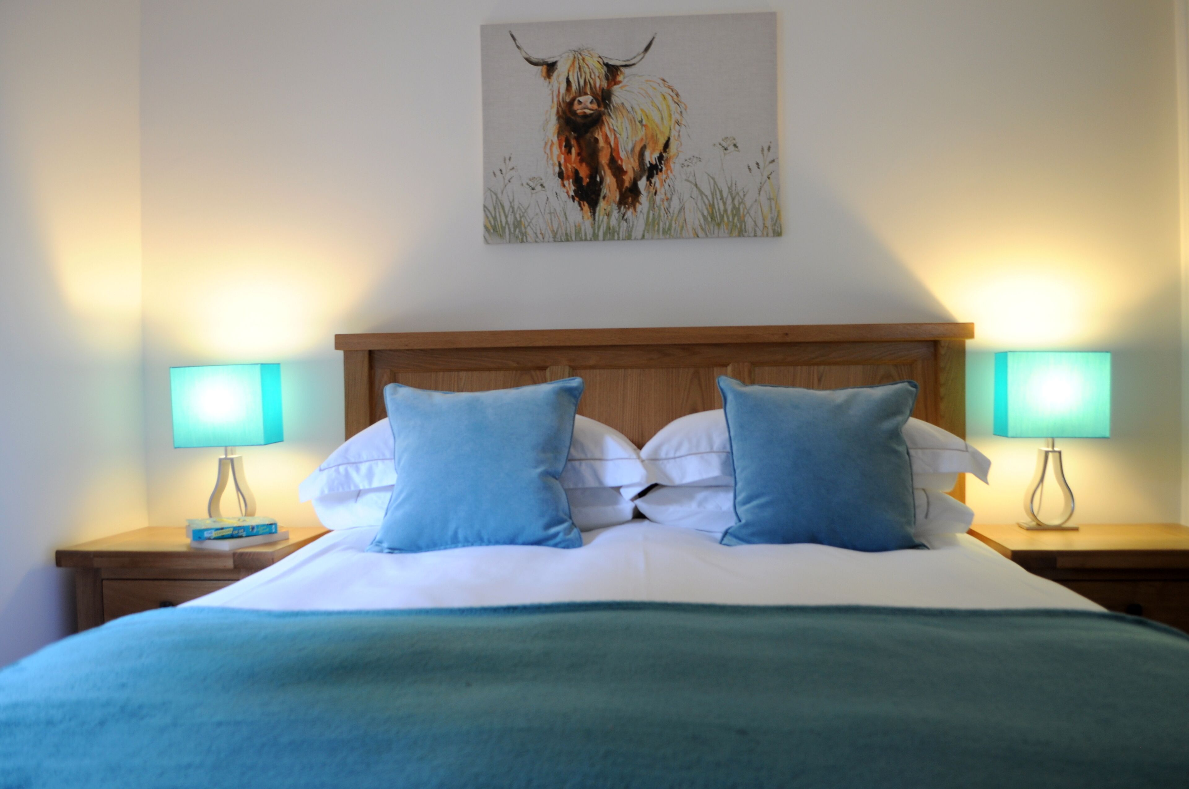Una cama bien hecha con almohadas azules y una manta verde, flanqueada por dos mesitas de noche con lámparas, y un cuadro de un toro en la pared.
