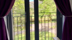 Ein Schlafzimmer mit einem Bett, violetten Vorhängen und einem Balkon mit Blick auf Grünflächen und eine Straße.