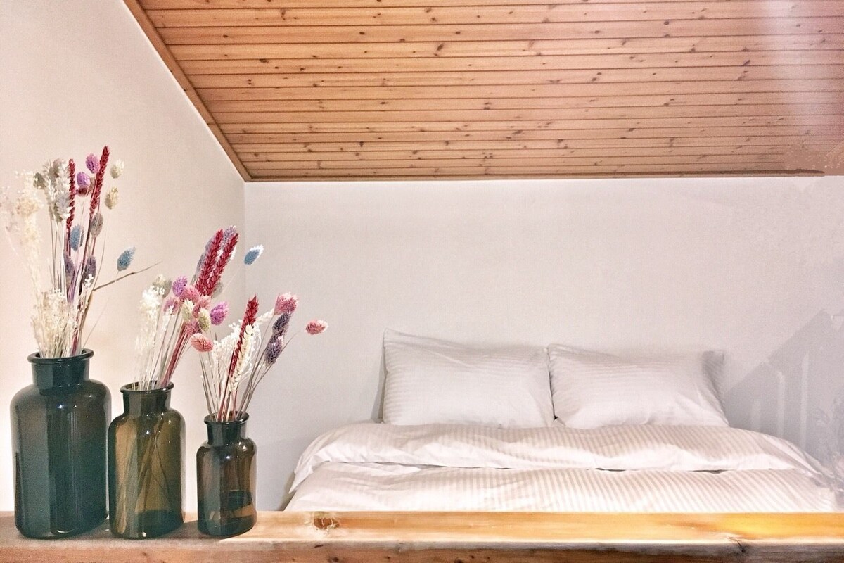A bedroom with a wooden ceiling, a bed with white bedding, and three glass vases with dried flowers on a wooden shelf.