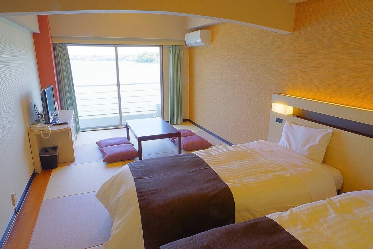 Een hotelkamer met twee bedden, een bureau, een stoel, een tafeltje en uitzicht op het omliggende landschap door een groot raam.
