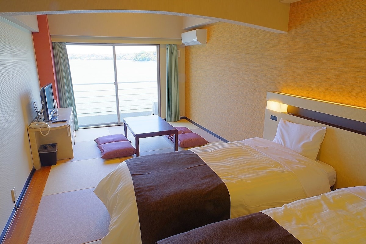 Een hotelkamer met twee bedden, een bureau, een stoel, een tafeltje en uitzicht op het omliggende landschap door een groot raam.