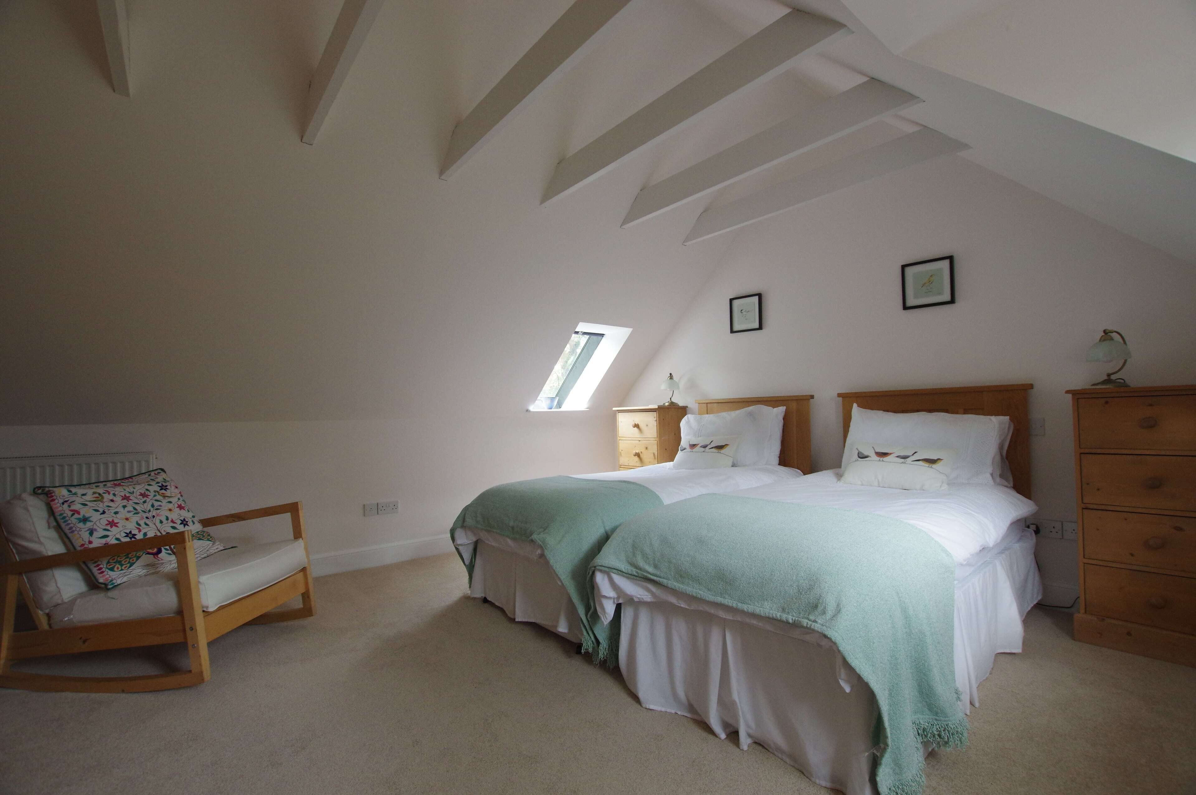 A bedroom with a sloped ceiling, a bed with a green blanket, a wooden rocking chair, and a dresser.