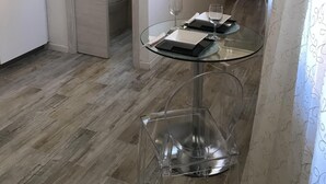 Ett litet matområde med ett glasbord och stolar, en vit dörr som leder till ett badrum och en säng med en röd kudde i bakgrunden.