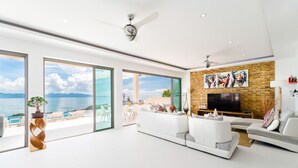 A spacious living room with a large white sofa, a flat-screen TV mounted on a brick wall, and a view of the sea through large windows. - Luxury 5-bedroom villa with panoramic sea view (Koh Samui)