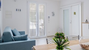 A modern living room with a sofa, a dining table with chairs, and a large sliding glass door leading to an outdoor area. - Vacation Home Offers, Salento, Gallipoli-Torre San Giovanni di Ugento (Posto Rosso Alliste Marina)
