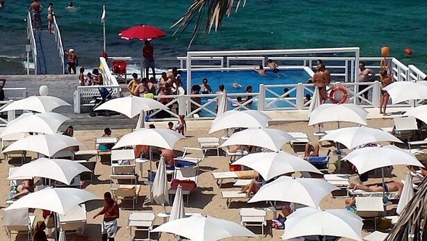 A beachside pool area with multiple white umbrellas and lounge chairs, surrounded by a railing and palm trees. - Vacation Home Offers, Salento, Gallipoli-Torre San Giovanni di Ugento (Posto Rosso Alliste Marina)