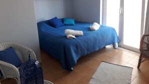 Ein Schlafzimmer mit Bett, Kissen, einem zusammengerollten Handtuch, einem Korbstuhl, einem kleinen Tisch und einem Gemälde an der Wand.