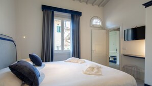 A hotel room with a bed, a TV, a window with curtains, and a door leading to another room.
