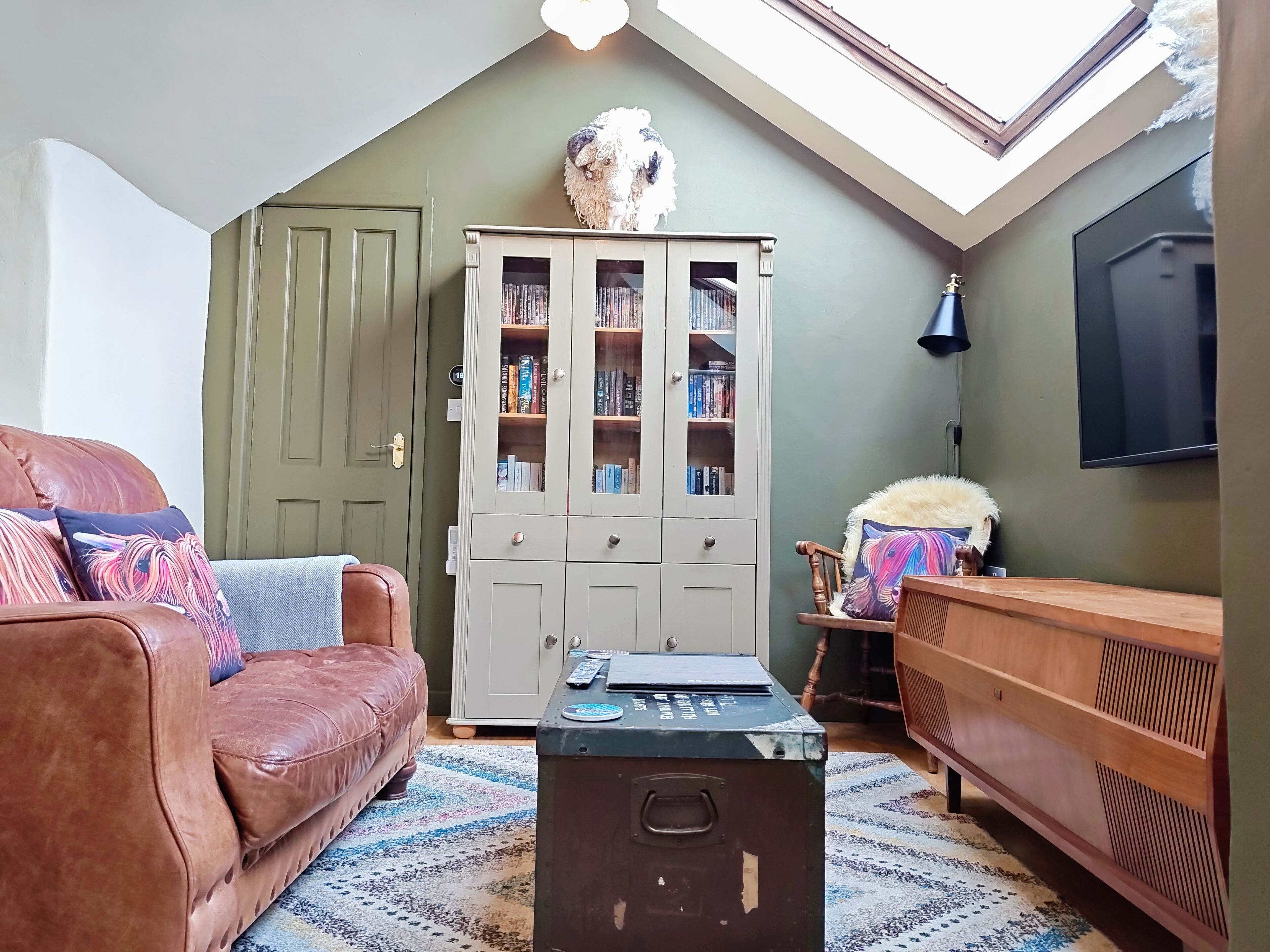 A cozy living room with a brown leather sofa, a wooden side table, a bookshelf, and a television mounted on the wall.