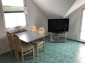 Un área de comedor con una mesa y sillas, un televisor sobre un pedestal de vidrio y una ventana con persianas.