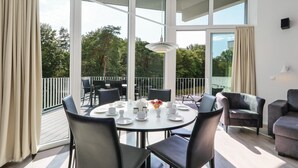 Ein moderner Essbereich mit einem runden Tisch, gedeckt für vier Personen, umgeben von Stühlen. Große Fenster bieten einen Ausblick ins Freie.