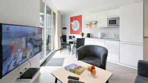 A modern living room with a flat-screen TV, a dining area with a table and chairs, a kitchen with white cabinets and stainless steel appliances, and a large window with curtains. - Dünenvilla 12, Whg 2 (Travemünde Waterfront)